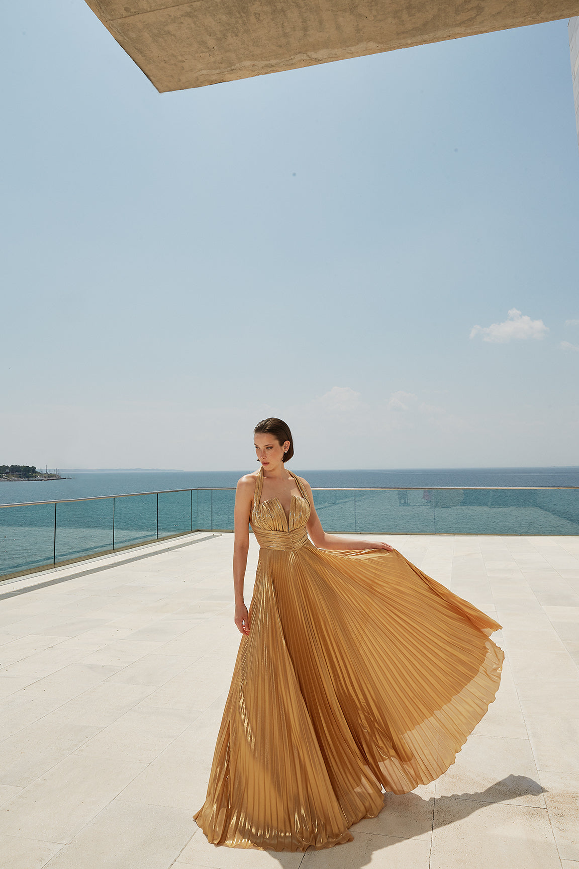 femme dans une longue robe debout sur un patio au bord de l’océan, robe dorée, élégante robe grecque antique, drapée d’or soyeux, robe fluide, déesse grecque, robe fluide