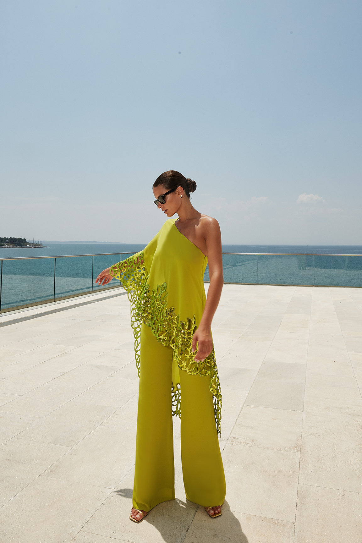 femme en robe jaune debout sur un patio près de l’océan, salopette jaune, soie fluo fluide, près de la mer