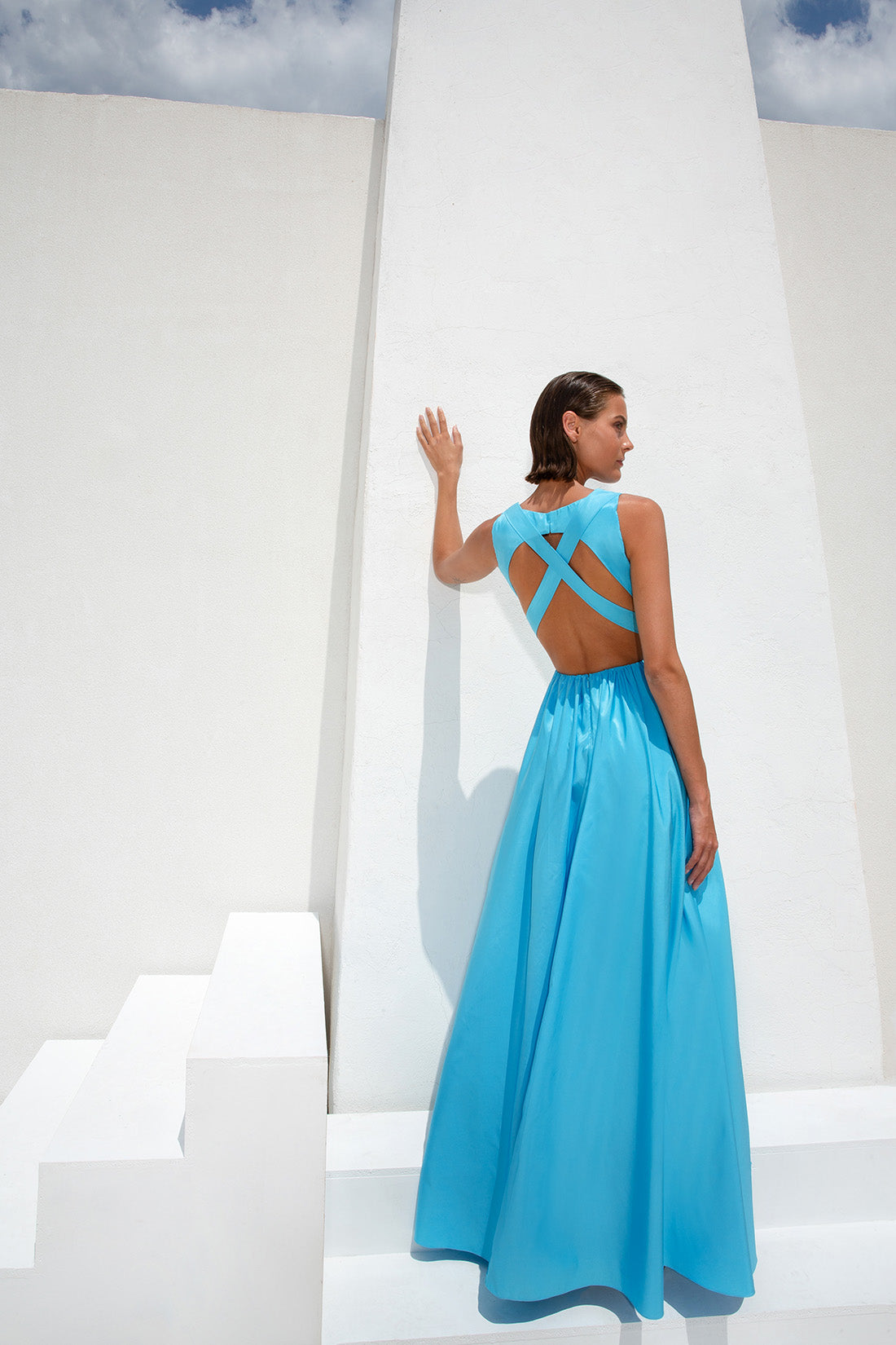 femme dans une robe bleue debout sur un mur blanc, longue robe bleue, robe bleu ciel, robe fluide, robe en soie bleue, robe dos nu, robe fluide, longue robe fluide luxueuse, robe bleue, robe longue