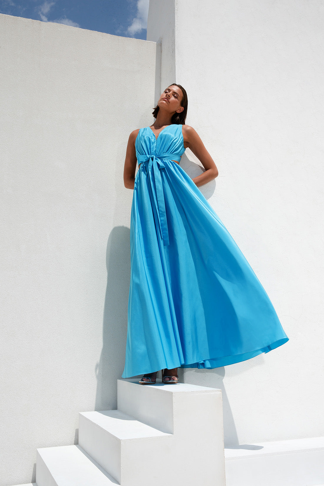 femme dans une robe bleue debout sur un mur blanc, longue robe bleue, robe bleu ciel, robe fluide, robe en soie bleue, robe dos nu, robe fluide, longue robe fluide luxueuse, robe bleue, robe longue